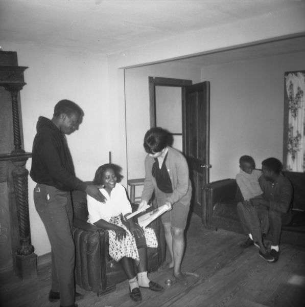 A woman and a man stand and talk to a seated woman. The standing woman is showing her papers. In the background, a man sits on a sofa talking to a child seated on his lap. "MFDP (Mississippi Freedom Democratic Party) workers talk to Mrs. Ruth Graves in Tibbee. Her daughter is attending the white school."