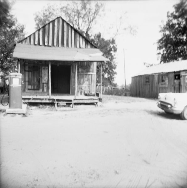 A small store with porch and a gas pump out in front. A car is parked nearby on the right. A man stands in the doorway of the neighboring building. "The store of Mr. Adell Bennett, candidate for the ASCS (Agricultural Stabilization and Conservation Service) for the second year. He lost both times."