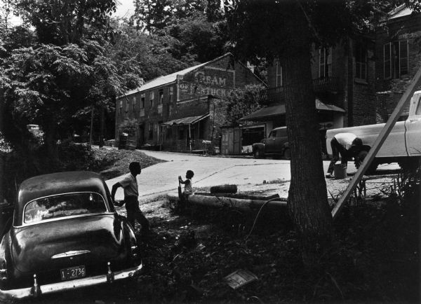 Several cars are parked outdoors. To the right, a man washes his pickup truck. To the left, a man leans against a car. Nearby, a child is seated on a short wall. In the background, residential and commercial buildings are visible.