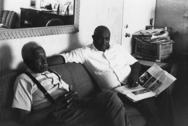 Two men sitting on a sofa. On the right is Amzie Moore, noted civil rights activist and entrepreneur, with a newspaper on his lap.