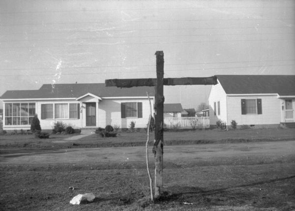 A charred wooden cross attached to a stake in the ground on the lawn near a road. In the background are residential buildings.