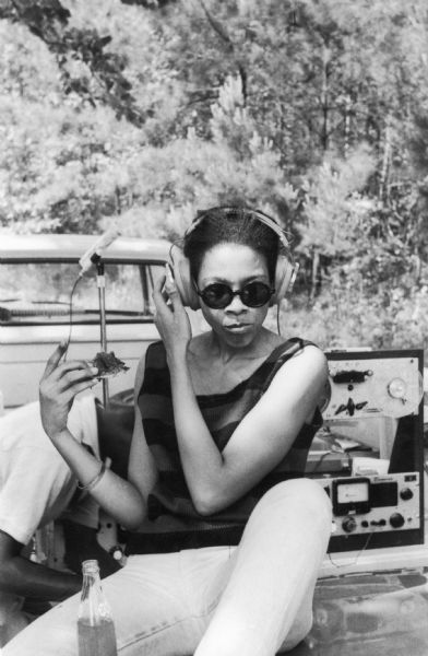 A woman, identified as Tracy, friend of Rob McNamara and supporter of SNCC (Student Nonviolent Coordinating Committee), wears headphones and eats. Behind her, a man bends over reel-to-reel audio equipment.<p>This photograph is tentatively identified as taken at a barbeque hosted by Mr. Steptoc for SNCC staff in McComb, Mississippi.