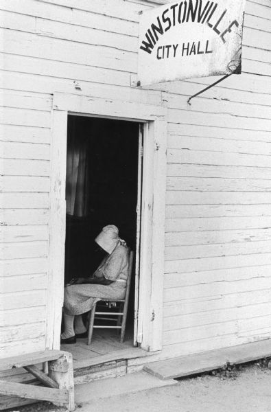 A woman wearing a bonnet sits inside the open doorway of a building. A sign above the doorway reads "Winstonville City Hall."
