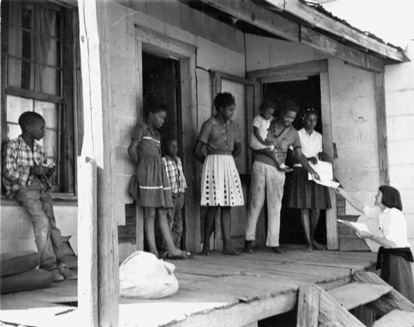A Caucasian woman, identified as Barbara Deming, hands leaflets to an African American family on a front porch. The location is tentatively identified as Georgia. This image comes from the collection of Carl and Anne Braden, who were both civil rights activists with SCEF (Southern Conference Educational Fund).