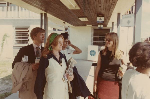 Three travelers stand at the pickup location for gate 4 at the Barbados Airport. On the back of the photograph they are identified as "Jim Hoge (editor of the "Chicago Sun Times"), Alice Hoge (now Alice Arlen), & Frankie Fitzgerald." The are attended by two women who appear to be airport employees.