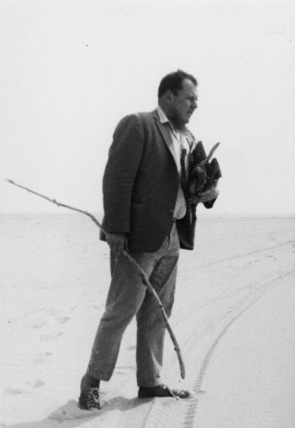 Emile de Antonio stands on a beach wearing a suit coat, pants and shoes while carrying logs and sticks of different sizes. He is turned to the side and is looking off to the right. The back of the image reads "Beach at East Hampton 1971 - Wood for picnic."
