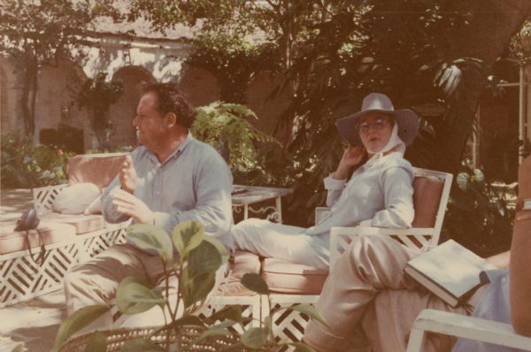 Three people sit in pink and white lounge chairs on a large outdoor patio surrounded by green plants and trees. Emile de Antonio sits next to socialite Marietta Tree. De Antonio looks off to the left, while Tree looks at the camera. On the right can be seen the legs and left arm of an unidentified person seated with a book in their lap. The patio is dappled by sunlight streaming through the foliage. The back reads "Marietta Tree and I - Barbados 1959."