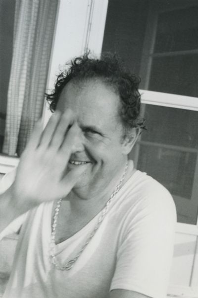 Waist-up seated portrait of De Antonio sitting in front of a door. He is smiling and holding a blurry hand up to the camera, obscuring part of his face. Dressed very casually, he wears a plain cotton t-shirt, and a chain around his neck. The back reads "Summer 74 - East Hampton."