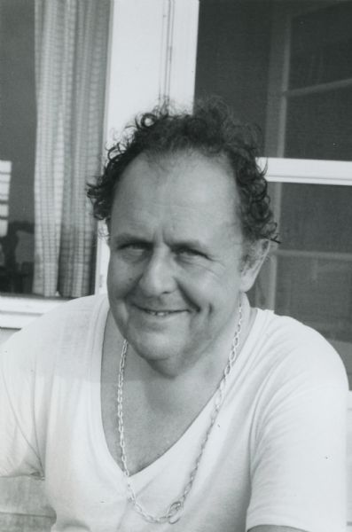 Waist-up seated portrait of De Antonio, who is smiling and seated in front of a door. Dressed very casually, he wears a plain cotton t-shirt, and a chain around his neck. "Summer 74 - East Hampton."