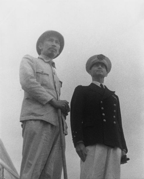 Low angle three-quarter length view of Ho Chi Minh standing with Admiral D'Argenlieu. Both men are in uniform.
