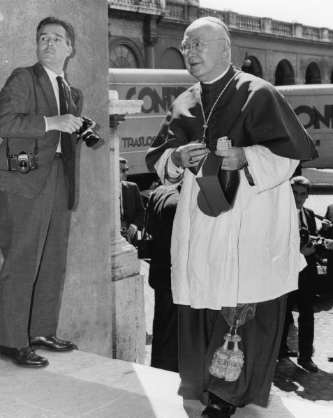 Cardinal Spellman stands with hat in hand looking off to his right. His full body and Cardinal robes are visible. In the left third of the image, a photographer stands to the side of the Cardinal, camera in hand, but looking off to the right, grimacing. In the background, moving vans and and camera men fill a corner of the Vatican complex.