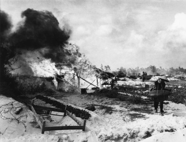 Typed note on the back: SGP...UNIPIX  North of Hue, South Vietnam: A Vietnamese woman carries her possessions as her home burns behind her. The South Vietnamese ARVN 1st Infatry moved more than 4,500 civilians from a village north of Hue and burned their homes to clear the area for a "Free fire zone."