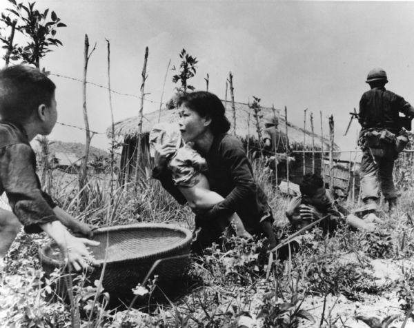 A woman is placing a baby in a basket held by a little boy. Behind her, a man lies prone holding another child, while two soldiers walk towards a house in the background. Behind them is a fence and beyond are thatched roof dwellings.