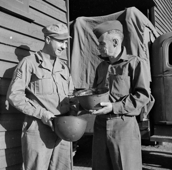 Sergeant William (Hub) Gilligan of Camp Douglas, Wisconsin and Sergeant Elmer Warnke of Pardeeville, Wisconsin, conversing at the Quarter Master warehouse at Logan Village located at Camp Cable, near Brisbane, Australia. They are both holding their helmets.