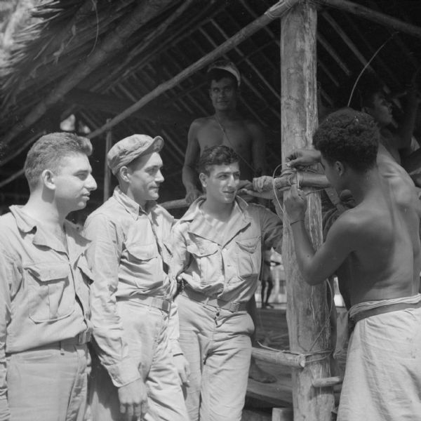 Three soldiers from the U.S. 6th Army Division, from Detroit, (left to right), Private Ned Zaffaramo, Private First Class Richard Rheaume and Private Kamil Abdo. They are talking to an indigenous man named Krasia as he ties a knot holding up a railing on a building located on Milne Bay, New Guinea (present day Papua New Guinea).
