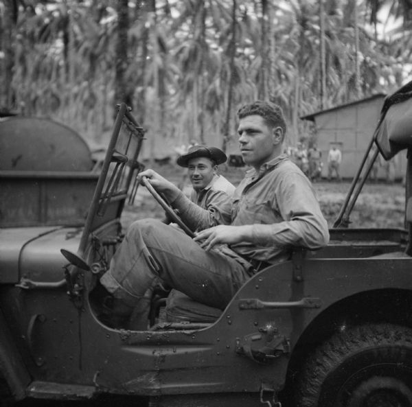Two soldiers pose in a jeep. The driver is Sergeant Howard Cody of Racine, Wisconsin, and next to him is Private Eugene Pregont of Wausau, Wisconsin. Palm trees, buildings and several men are in the background. The medical unit camp area is located on Milne Bay, New Guinea (present day Papua New Guinea).