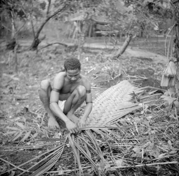 An indigenous youth weaves roofing for military buildings on Kiriwina Island in the Solomon Sea, New Guinea (present day Papua New Guinea).