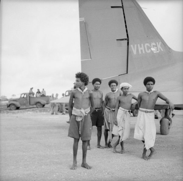 Indigenous men stand by a tail section as they watch planes take off and land at the Kiriwina Airfield on Kiriwina Island in the Solomon Sea, New Guinea (present day Papua New Guinea). Soldiers and trucks are visible in the background.