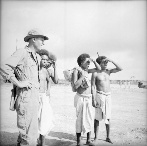 A soldier and four indigenous men shade their eyes as they watch planes take off and land at the Kiriwina Airfield on Kiriwina Island in the Solomon Sea, New Guinea (present day Papua New Guinea). The soldier has a rifle slung over his shoulder.