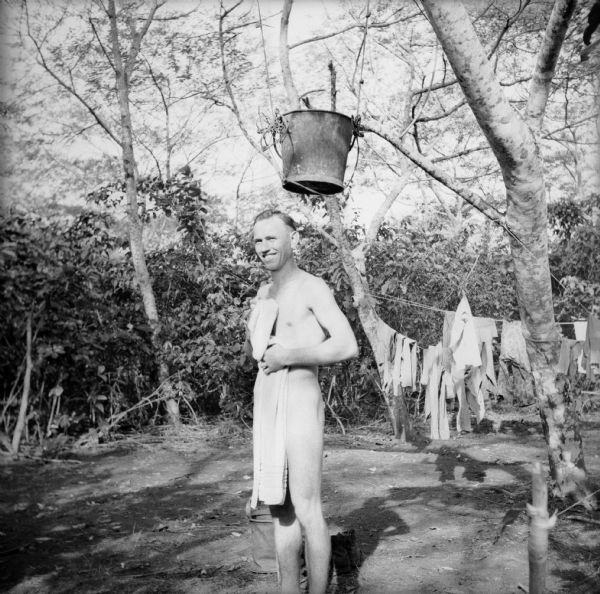Private Clarence Charneski, Surgical Technician-Medical Detachment, of Green Bay, Wisconsin, takes an improvised outdoor shower under a bucket at a military camp on Kiriwina Island in the Solomon Sea, New Guinea (present day Papua New Guinea). He was drying off when the photo was snapped.