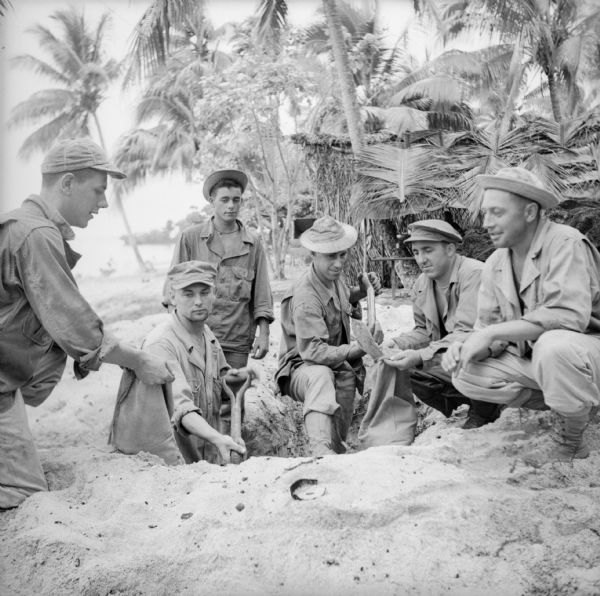 Soldiers working on slit trench in the rain on Kiriwina Island in the Solomon Sea, New Guinea (present day Papua New Guinea). They all hail from Michigan. Left to right, Private Edward Glomski of Hazel Park, Private 1st Class Ed. Abbott of Saginaw, Private Kenneth Steele of Saginaw, Corporal Neil Story of Clio, Corporal Eugene Drury of Hazel Park and Corporal John Blohm of Saginaw.