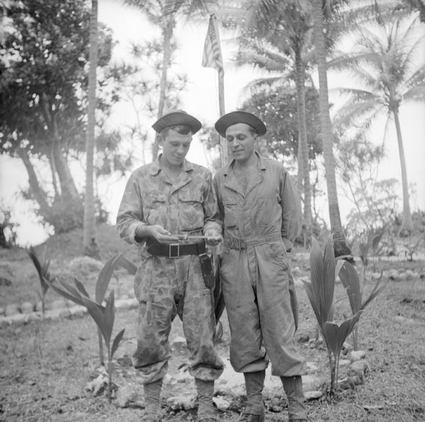 Two soldiers inspect a knife at Kiriwina Beach Camp on Kiriwina Island in the Solomon Sea, New Guinea (present day Papua New Guinea). They are standing in front of a flagpole in a garden, flying the American flag. On the left is Private Frank Pollen of Manitowoc, Wisconsin and on the right is Private Herman Oxendorf of Milwaukee, Wisconsin.
