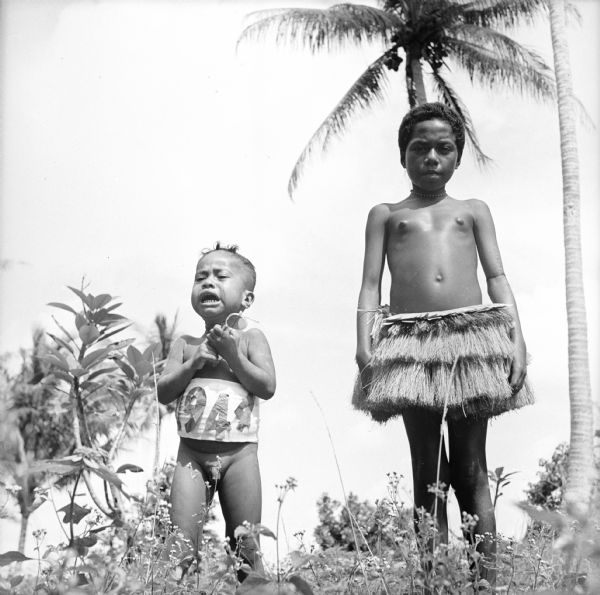 View looking up at an upset indigenous toddler wearing a banner around his middle with "1944" pasted on it. He is holding a pair of sunglasses and standing next to a young indigenous girl wearing a grass shirt. Image taken on Kiriwina Island in the Solomon Sea, New Guinea (present day Papua New Guinea).