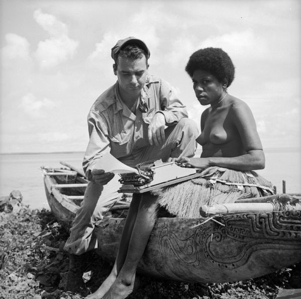 An indigenous woman wearing a grass shirt sits on a carved boat with Robert Doyle on the shore of Kiriwina Island in the Solomon Sea, New Guinea (present day Papua New Guinea). She is using Robert's typewriter and he is holding the emerging typewritten copy.