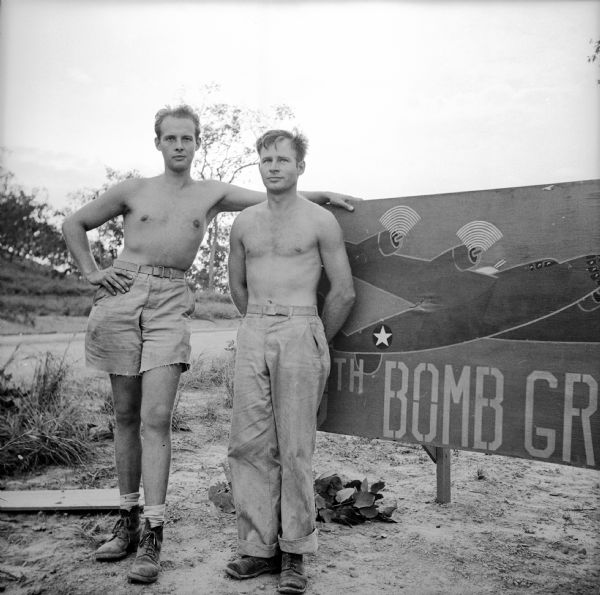 Two Wisconsin cousins, (left) Corporal Englund Johnson, bomber waist gunner, of Park Falls and (right) Sergeant Donald Englund, airplane engine mechanic, of Prentice, are posed shirtless in front of a painted sign at the Kiriwina Airfield on Kiriwina Island, in the Solomon Sea, New Guinea (present day Papua New Guinea). They reunited for the first time in three years on March 16th, 1942, in Darwin, Australia. The sign probably reads, "90th Bomb Group." In Doyle's notes, Sergeant Englund is with the "319th Sq. 90th Bomb."