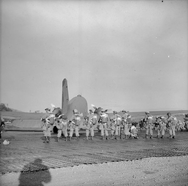 Paratroopers at a Drome (airfield) near Port Moresby, preparing to take off for a landing at Nadzab near Lae. Lae is a Japanese-held airfield in New Guinea (present day Papua New Guinea). The airplane is behind the group. The shadow of Robert Doyle taking the photograph is in the lower left corner.