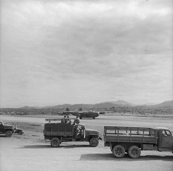 Trucks at a Drome (airfield) near Port Moresby, wait while paratroopers prepare to take off for a landing at Nadzab near Lae. Lae is a Japanese-held airfield in New Guinea (present day Papua New Guinea). There is an airplane on the runway.