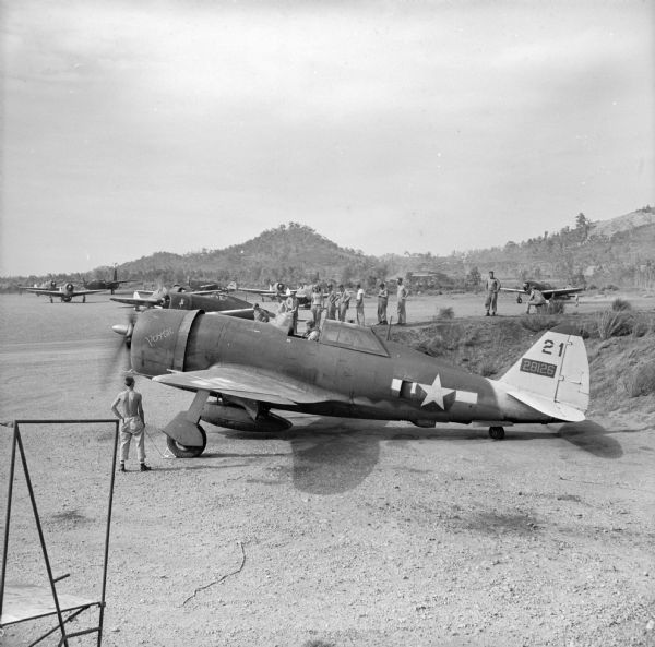 Military aircraft at a Drome (airfield) near Port Moresby, preparing to take off for a landing at Nadzab near Lae. The aircraft in the foreground is named "POYSE" and the numbers on the tail are "21, 28126." Many soldiers are watching. Lae is a Japanese-held airfield in New Guinea (present day Papua New Guinea).