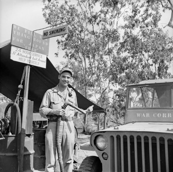 A smiling Robert Doyle holds the nozzle to the gas pump with one hand and gives the "thumbs up" sign with the other at the Kangaroo Korner Gas Station near the entrance to the 5th Air Force Headquarters near Port Moresby, New Guinea (present day Papua New Guinea). He is filling his jeep with fuel. "War Corr[espondent]" is stenciled below the windshield. Signs overhead read, "We Fill Anything Bound For Japan For 'Free,'" "24 Hour Service, If Attendant Isn't Around, Help Yourself" and "No Smoking." Behind Doyle is a tent. On the back of the contact print is written, "Here's Where Your Gas Is."