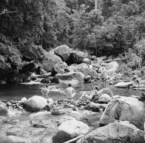 Robert Doyle's caption says it best, "The ol' swimmin' hole, South Pacific style. Every afternoon it is filled with shouting soldiers in their birthday suits, seeking relief from the sweltering heat, swimming in the cold, crystal clear water or lounging on the huge rocks. The natural pool, formed by a cascading mountain stream, is 12 feet deep." In another caption he says, "One compensation for the tropical heat, dust, mud and insects is this mountain pool discovered by our soldiers." The soldiers uniforms can be seen drying on the rocks. The swimming hole is located on Goodenough Island, in the Solomon Sea, New Guinea (present day Papua New Guinea).