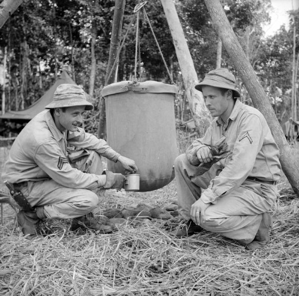 Twin soldiers, Corporal John Kellner (left) and Private First Class Bob Kellner (right) of Milwaukee get a drink at the lister bag, in the camp on Goodenough Island, in the Solomon Sea, New Guinea (present day Papua New Guinea). Corporal Kellner is wearing a knife and both soldiers are wearing and holding gloves. Trees and a tent are in the background. A lister bag (also spelled lyster bag) is a device used to disinfect drinking water.