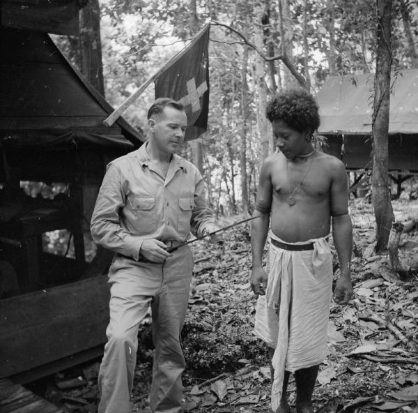 Major Austin Henry of Milwaukee, Wisconsin, poses with an indigenous man in front of the medical tent at the military camp on Woodlark Island, in the Solomon Sea, New Guinea (present day Papua New Guinea). They appear to be looking at a machete in the Major's hands. Trees and a tent are in the background.
