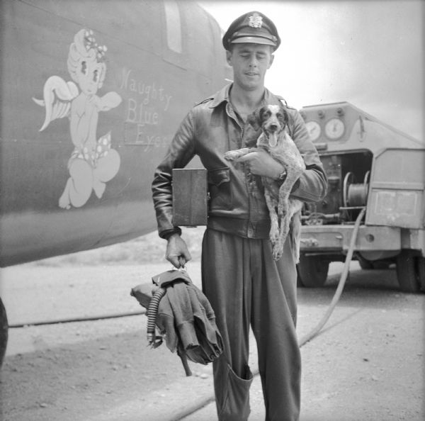 Navigator Lieutenant Paul Seramur of Milwaukee, Wisconsin, poses in front of a Consolidated B-24 Liberator warplane after the Alexishafen strike. The name and mascot of the plane, "Naughty Blue Eyes," is painted on the side of the warplane. Seramur is holding his pup, "Cobber" in one arm and his gear in the other. Alexishafen was a Japanese occupied airbase located on the northeast coast of New Guinea (present day Papua New Guinea), near Madang.