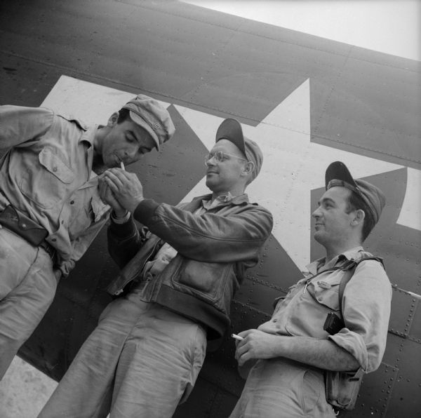 View looking up at three war correspondents, (left to right), Robert Doyle of the <i>Milwaukee Journal</i>, Bob Eunson of Associated Press and Ralph Boyce of <i>YANK</i> Army Weekly magazine take a smoking break in front of a warplane fuselage at Wards Drome (5 Mile), an airfield near Port Moresby, New Guinea (present day Papua New Guinea).