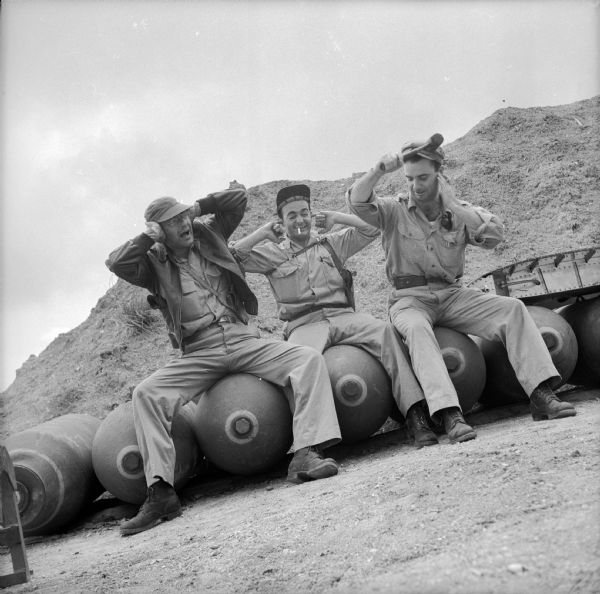 Three war correspondents, (left to right), Bob Eunson of Associated Press, Ralph Boyce of <i>YANK</i> Army Weekly magazine and Robert Doyle of the <i>Milwaukee Journal</i>, joke around at Wards Drome (5 Mile), an airfield near Port Moresby, New Guinea (present day Papua New Guinea). They are sitting on what appear to be bombs and are plugging their ears, waiting for the explosion. Doyle is also holding a sledge hammer to his head. Robert Doyle notes that this photograph was taken by Sergeant Dick Hanley of <i>YANK</i>, the Army Weekly magazine, with Doyle's camera.