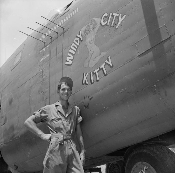 Mechanic Sergeant Raymond Smith of Ontario, Wisconsin, stands next to the nose art, "Windy City Kitty," depicting a naked woman in the "pin-up style" on the fuselage of his warplane. The airfield is near Port Moresby, New Guinea (present day Papua New Guinea). Robert Doyle wrote a caption for this image although it was not published at that time, "Airman poses beside B-24 at airstrip in New Guinea. Plane is named after cartoon character featured in <i>Yank Down Under</i>, Australian edition of Army Magazine <i>Yank</i>." She was a "racy cartoon character named 'Windy City Kitty,' from Chicago, Illinois," created by Sergeant Jack Crowe.

