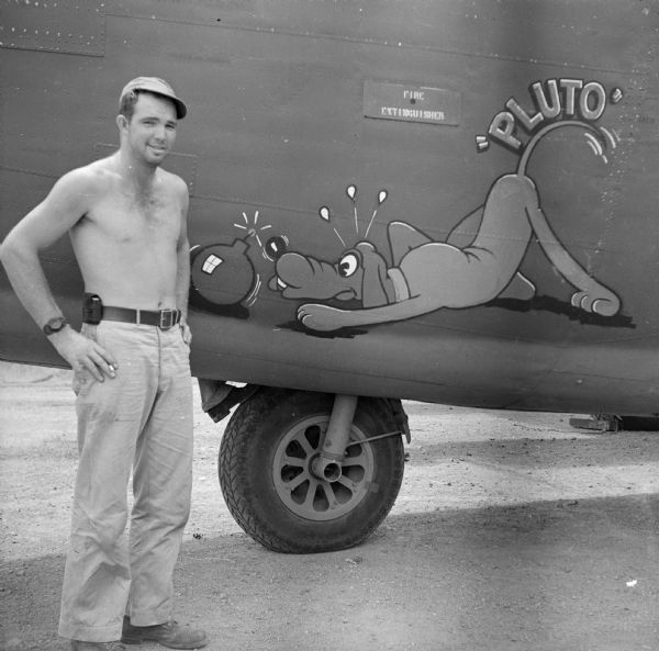 Mechanic Staff Sergeant Raymond Wagner of Boscobel, Wisconsin, poses shirtless while holding a cigarette and standing next to the nose art on his Consolidated B-24 Liberator warplane. In the design, Mickey Mouse's cartoon dog "Pluto" is sniffing a bomb. The airfield is located near Port Moresby, New Guinea (present day Papua New Guinea).