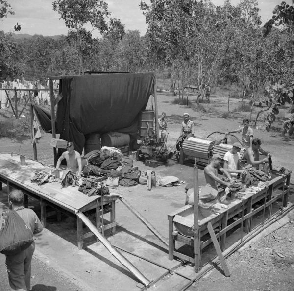 Elevated view of the Rough Raider Squadron Laundry, serving 100 customers a day. Eight soldiers can be seen, some working, some watching, and one soldier carrying a bulging laundry bag over his shoulder. Trees are  in the background, and drying garments are on the left. The laundry is located near Port Moresby, New Guinea (present day Papua New Guinea).