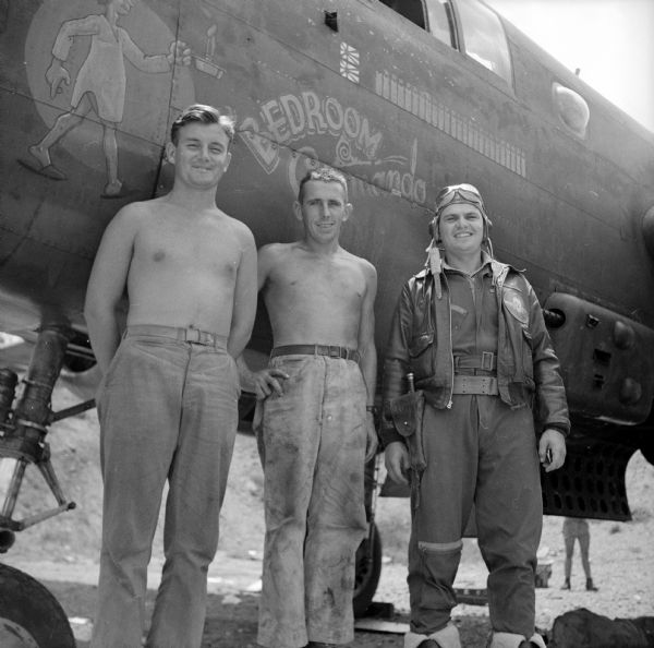 Three Wisconsin soldiers pose in front of the nose art on their warplane, "Bedroom Commando," depicting a sleepwalker in a nightshirt and slippers, carrying a candle. Left to right are Crew Chief Sergeant Clyde Cheney (shirtless) of Milwaukee, Instrument Specialist Sergeant Leander A. Grosse (shirtless) of Cross Plains and Navigator and Bombardier Captain Sylvester Hoffman of Madison. Hoffman is wearing a leather bomber jacket, flying helmet and goggles. On the jacket is a patch with a skull and wings. The airfield is near Port Moresby, New Guinea (present day Papua New Guinea).