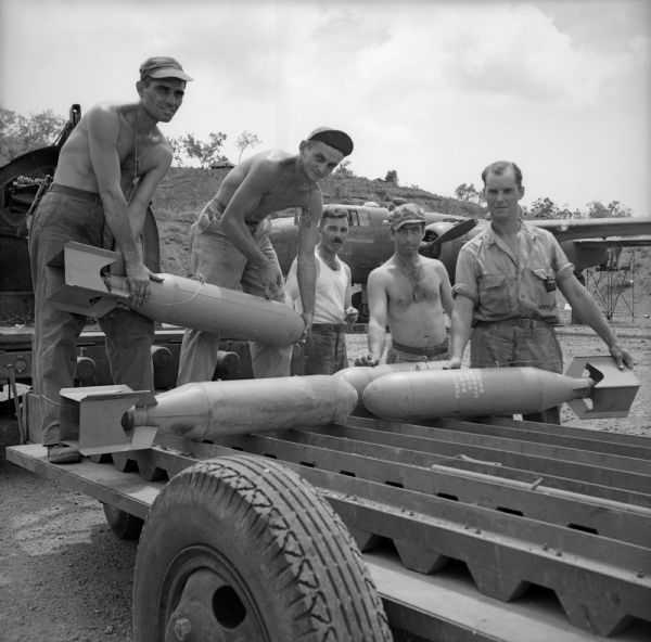 Michigan soldiers loading bombs onto a trailer at a military base near Port Moresby, New Guinea (present day Papua New Guinea). Their names, left to right, are Sergeant Joe Sankowski (shirtless) of Detroit, Sergeant Charles Sekula (shirtless) of Flint, Sergeant Leonard Welch of Detroit, Sergeant Edward Rushman (shirtless) of Wyandotte, and Sergeant Herman Rollin of Detroit. A warplane is in the background.