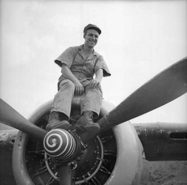 Armament Chief Sergeant Lloyd Cleary sits on the nose of a warplane with his feet on the propeller at an airfield near Port Moresby, New Guinea (present day Papua New Guinea).
