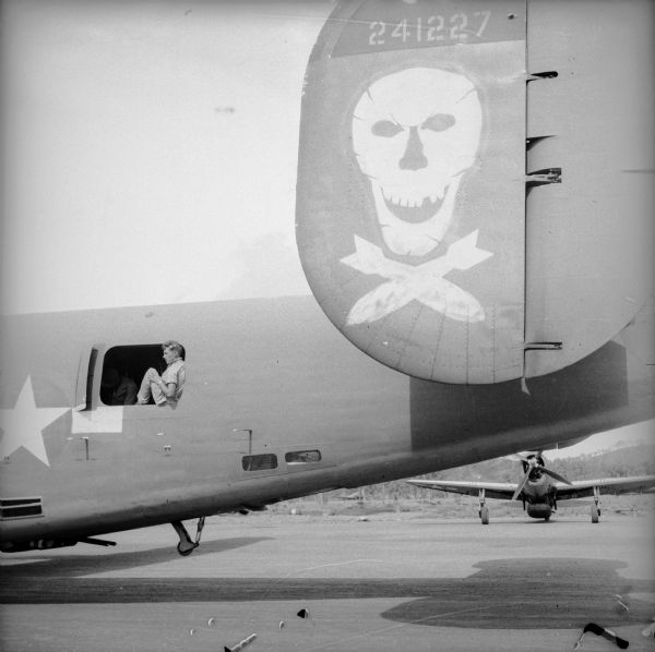 Skull and bombs tail art on a Consolidated B-24 Liberator warplane at New Guinea Airfield near Port Moresby, New Guinea (present day Papua New Guinea). A soldier is sitting in the gun door. A Republic P-47 Thunderbolt can be seen in the background.