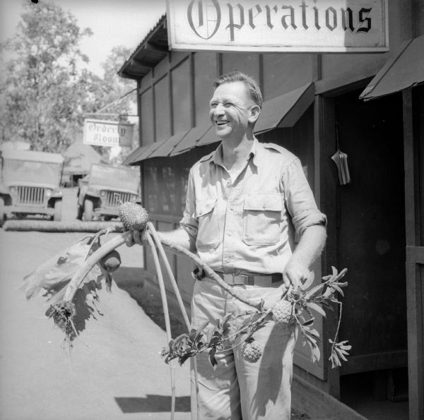 An Intelligence Officer smiles as he holds an indigenous plant with fruit on it used for food. A building is behind him with two signs, "Operations" and "Orderly Room." Two jeeps are parked in the background. The base is located near Port Moresby, New Guinea (present day Papua New Guinea).
