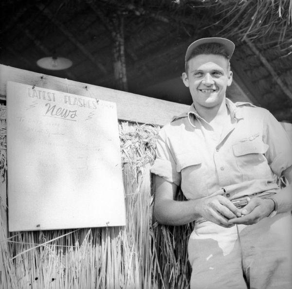 Lieutenant Clarence "Herk" Blend of Milwaukee, Wisconsin, smiles as he poses next to a bulletin board, "Latest Flashes - News" that is hanging on the side of a building. He was the pilot of a Republic P-47 Thunderbolt that shot down a "Tony" (an Italian-designed Japanese fighter aircraft) while escorting several Consolidated B-24 Liberator aircraft over Wewak, New Guinea (present day Papua New Guinea), the previous day.