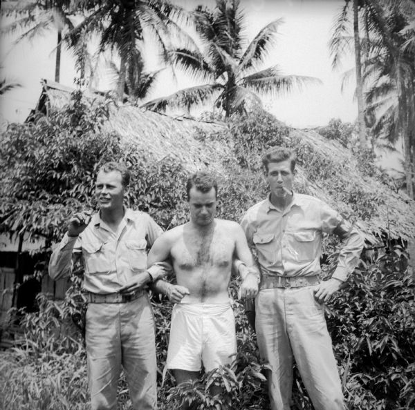 Robert Doyle poses in his undershorts between Private Gerald Minkin of Ironwood, Michigan (left) and Private First Class Samuel "Slim" Lanham of Louisville, Kentucky (right). Both soldiers are smoking cigars. A thatched roofed building and palm trees are behind them. The location is Gili Gili, a village, on Milne Bay, New Guinea (present day Papua New Guinea).