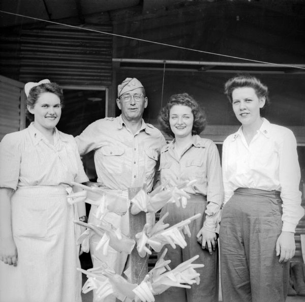 Three Wisconsin women and a man pose in front of a building. In the foreground is a rack holding rubber gloves. Names, left to right, are Lieutenant Kathleen Weckenmueller (in nurse's uniform) of Milwaukee, Lieutenant Colonel Leonard W. Peterson of Sun Prairie, Lieutenant Mildred Wehrwein of Milwaukee and Mary Blaschke, ARC, of LaCrosse (Catholic Welfare Bureau, Wausau).